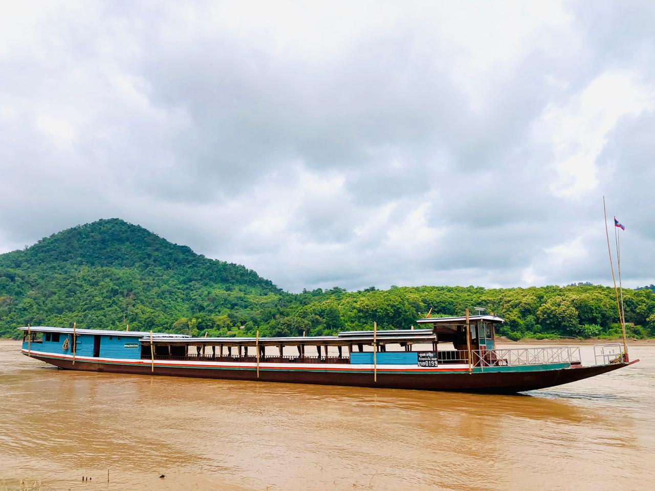 Long blue slow boat on the Mekong, forested hill behind it