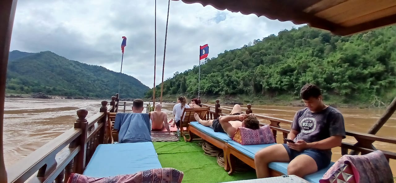 Passengers on deck mats, Lao flag, forested bank